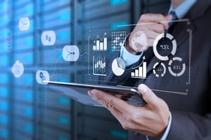 Geschäftsperson hält Tablet, im Vordergrund digitale Grafiken mit Diagrammen, Prozentangaben und Charts. Hintergrund wie Rechenzentrum oder Serverraum. Symbol für Datenanalyse, Technologie und Business-Management.
