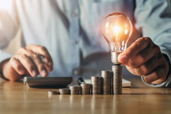 Hand hält Glühbirne über Stapel von Münzen in aufsteigender Höhe, daneben ein Taschenrechner. Symbol für Innovation, finanzielles Wachstum und Investitionsstrategien