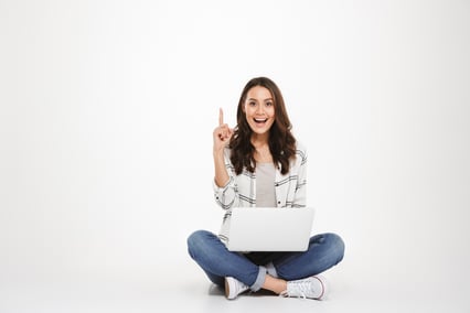 happy-brunette-woman-in-shirt-sitting-on-the-floor-with-laptop-computer-while-having-idea-and-looking-at-the-camera-over-gray