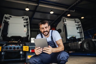 smiling-tattooed-bearded-blue-collar-worker-in-overalls-using-tablet-to-check-on-delivery-while-crouching-in-garage-of-import-and-export-firm